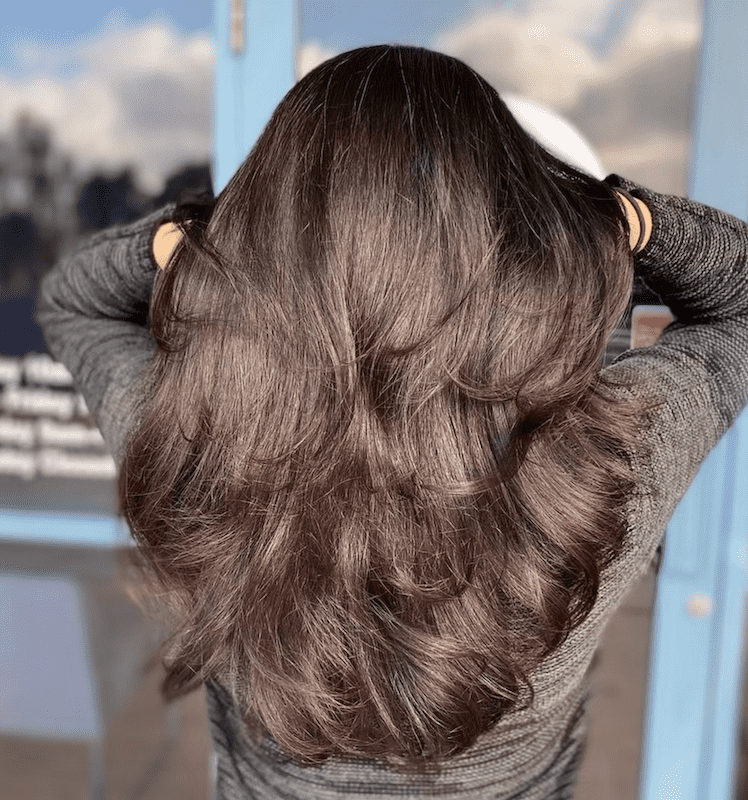 Long, wavy brown hair viewed from the back, hands adjusting hair outdoors.