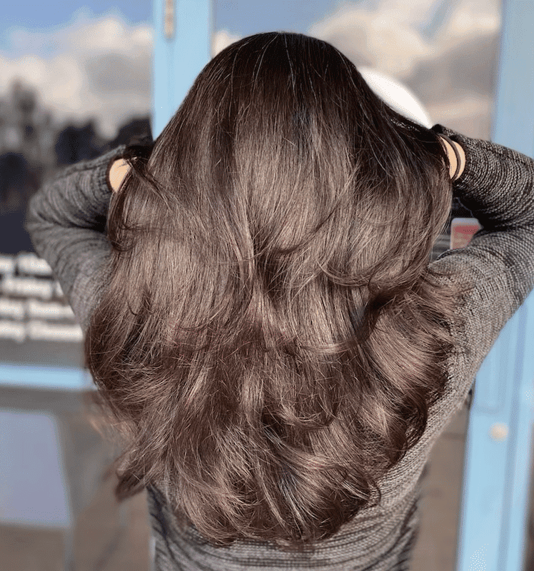 Long, wavy brown hair viewed from the back, hands adjusting hair outdoors.