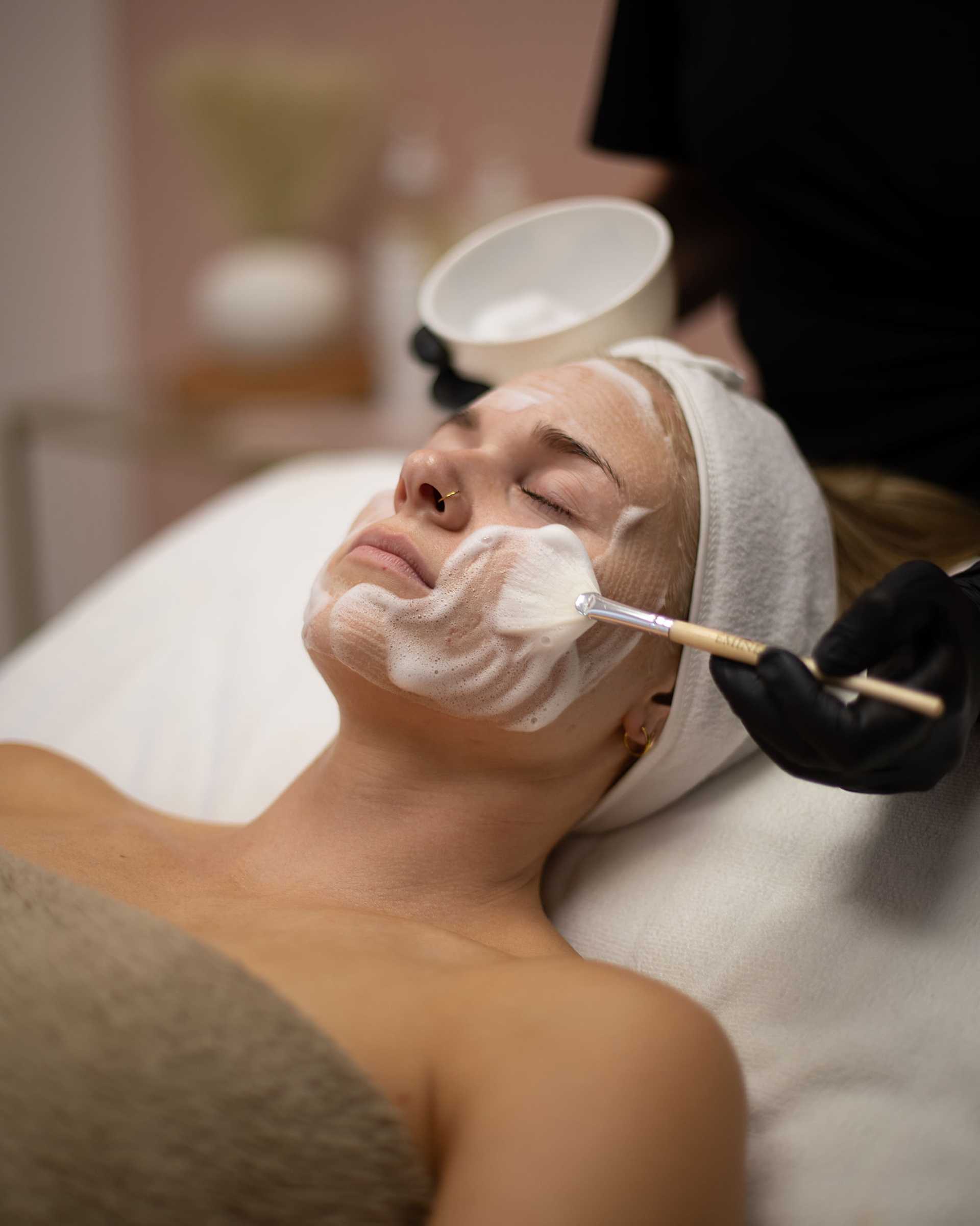 Person receiving a facial treatment with a brush and cream in a spa setting.
