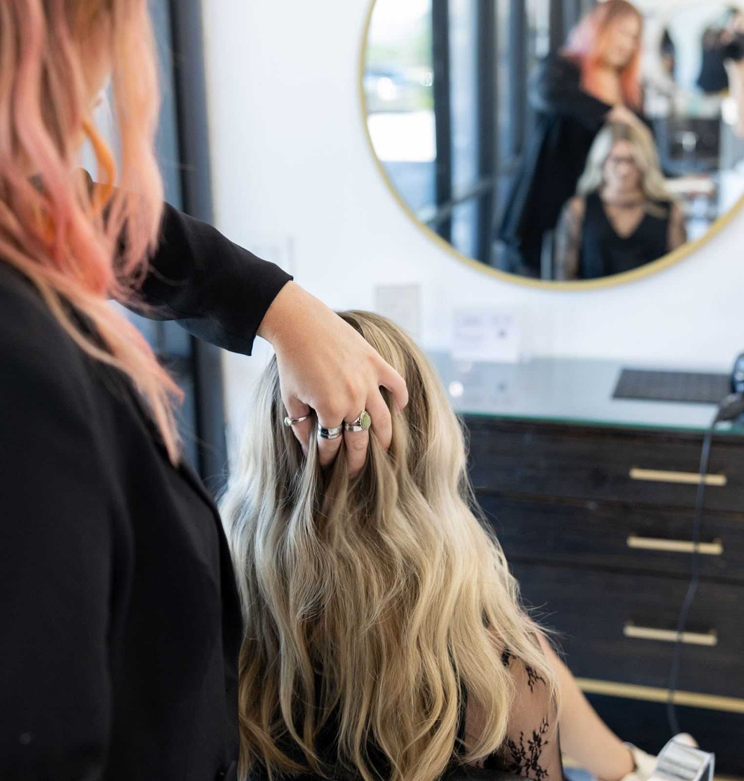 Hair stylist adjusting client's long, wavy blonde hair in a salon with a round mirror.