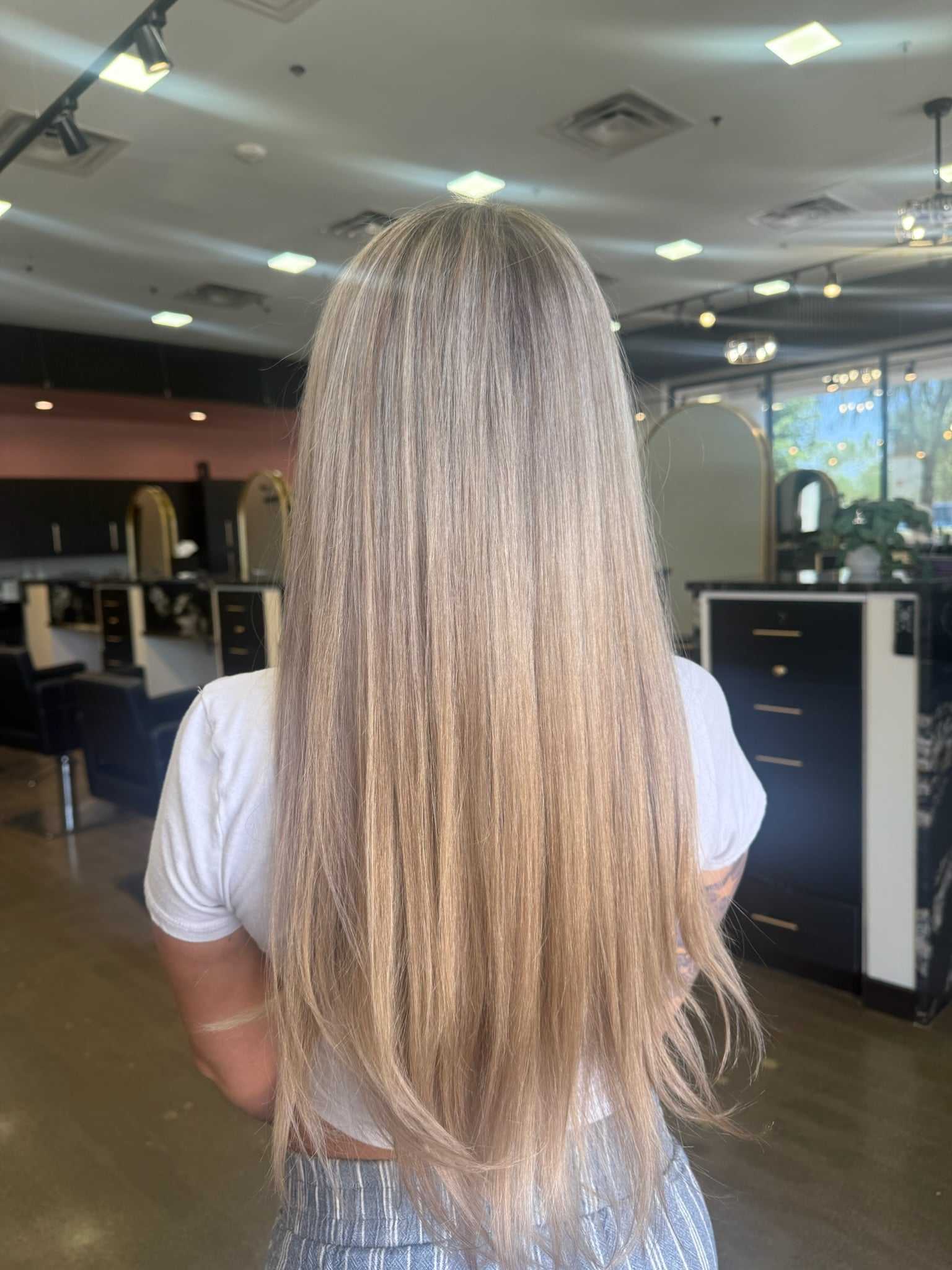 Woman with long, straight blonde hair in a salon, wearing a white shirt and striped pants.