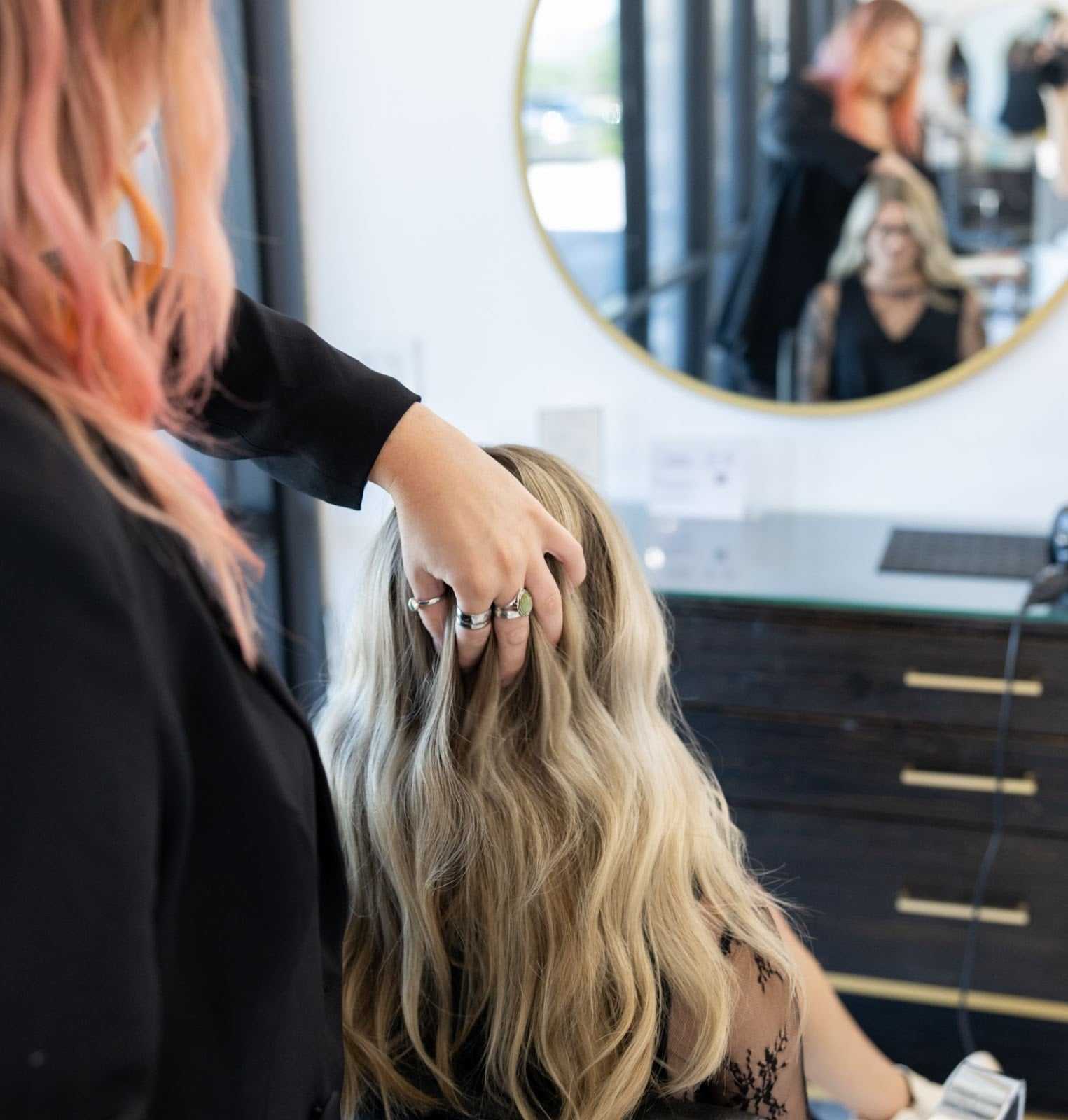 Hair stylist adjusting client's long, wavy blonde hair in a salon with a round mirror.